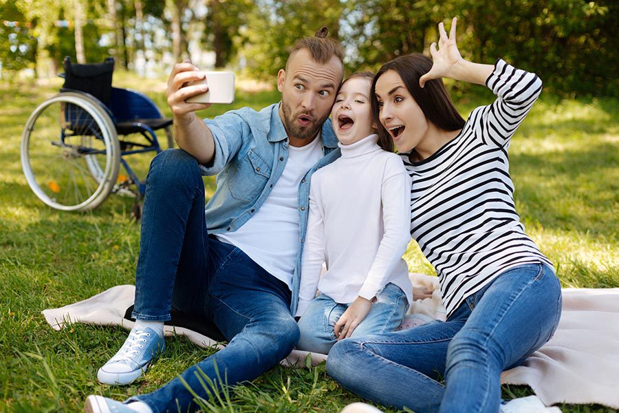 Personal Insurance - a Father, Daughter and Mother Take a Funny Selfie Photo, Sitting on a Picnic Blanket in a Park, a Wheelchair Sitting Behind Them