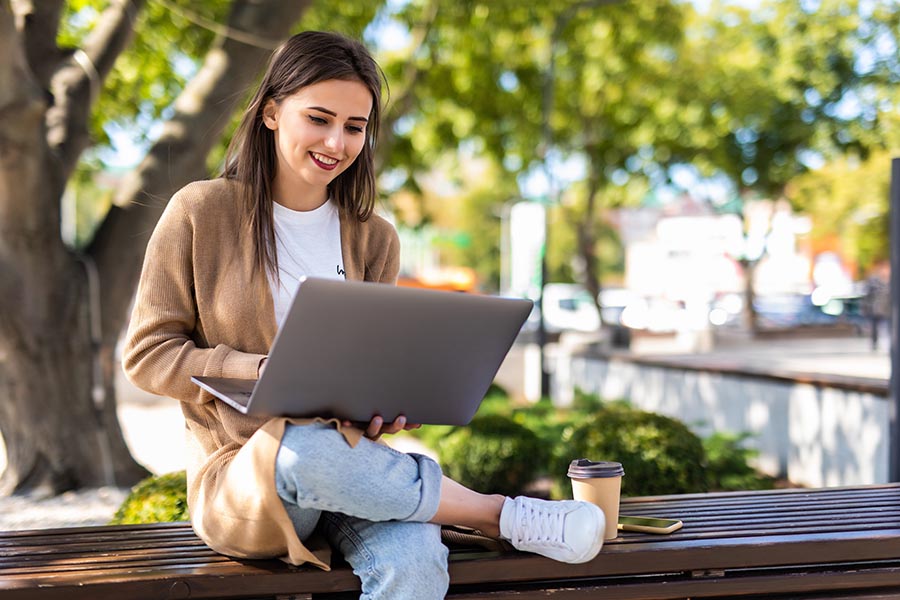 Blog - Young Woman Sits on a Bench in a Park and Reads on Her Computer, Smiling, With Legs Crossed
